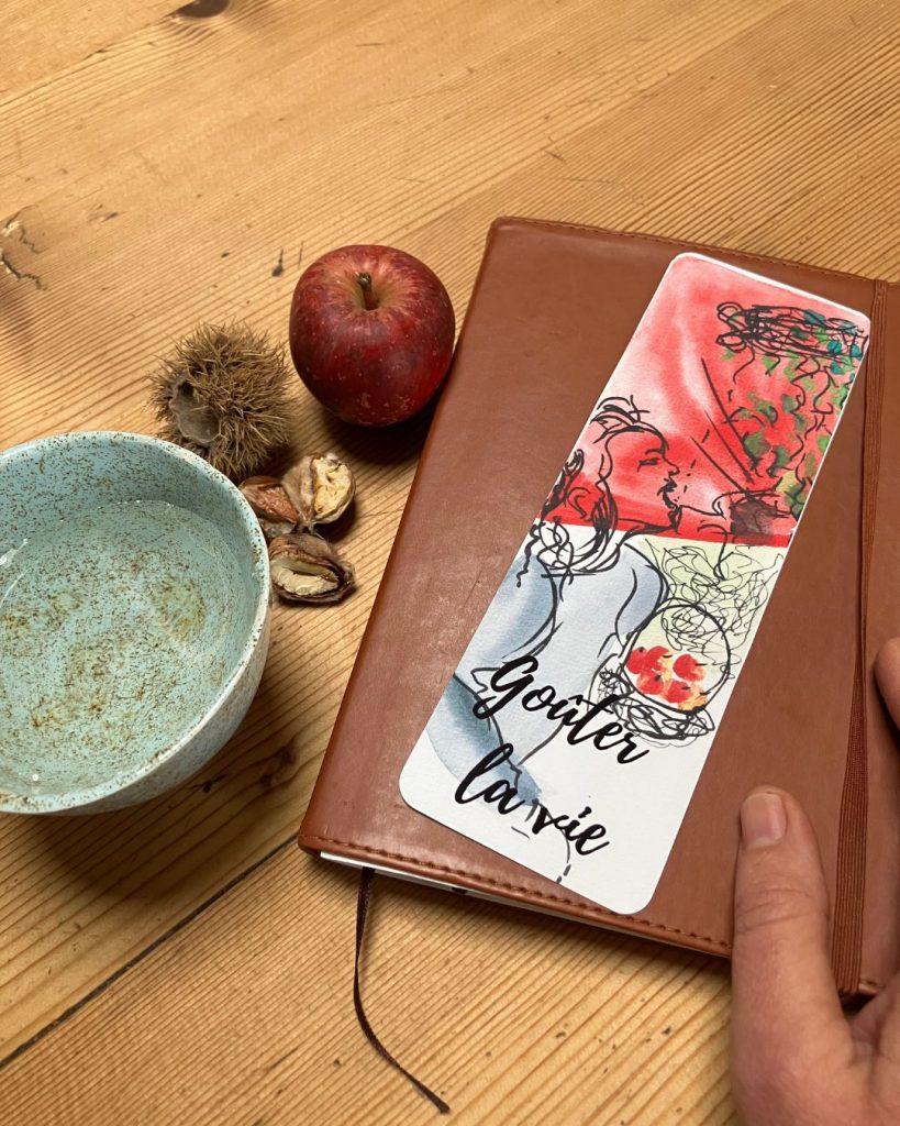 Le marque-pages "Goûter la vie" est vu du recto et posé sur un carnet fermé de couleur marron. Une main est posé sur le carnet, et à gauche du carnet on peut voir une pomme, une chataîgne et un bol de tisane.