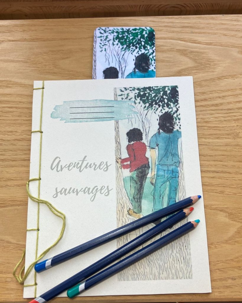 Carnet artisanal de la collection Enfants du Vivant fermé, où l'on voit la première de couverture, une illustration, accompagné d’un marque-page illustré, posé sur un support en bois.